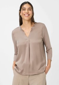 Donna sorridente con capelli scuri, indossa una blusa taupe con scollo a V e pantaloni beige, con le mani nelle tasche e una collana d'argento con due anelli.