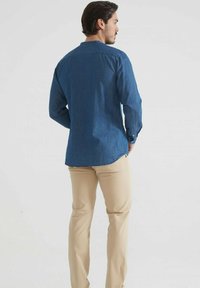 Homme portant une chemise à manches longues bleue et un pantalon beige, se tenant les mains dans les poches, tourné vers un fond uni.