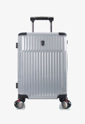 Valise rigide argentée avec des rainures verticales, quatre roues doubles pivotantes avec ressorts rouges, une poignée télescopique et un badge logo à l'avant.