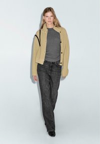 Cardigan beige a costine con bottoni neri, indossato sopra una maglietta grigia e jeans scuri, abbinato a calzature nere, mostrato su uno sfondo chiaro.