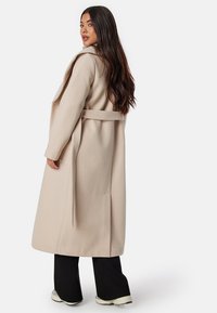 Beige dlouhý kabát s šálovým límcem, s páskem uvázaným v pase a bočními kapsami, znázorněný na modelu z zadního úhlu.