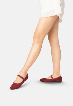 Jambes portant des ballerines Mary Jane en daim rouge vin avec des brides à boucle dorée, associées à une mini jupe blanche à volants, sur un fond uni clair.