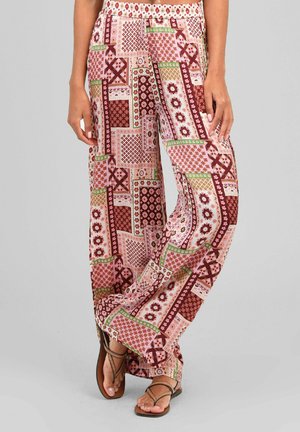 Femme portant un pantalon ample taille haute avec des motifs patchwork géométriques rouges, roses, verts et beiges et des sandales marron.