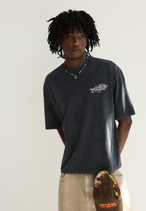 Man met dreadlocks die een donker grafisch t-shirt, beige broek, een kralenketting draagt en een bruine pet met gele en rode details vasthoudt.