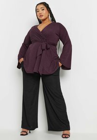 Top morado con escote en V y detalle de lazo, con mangas largas de campana. Combinado con pantalones anchos negros y sueltos.