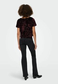 Īsām piedurknēm, bordo krāsas mirdzošs tops ar brīvu piegriezumu, savienots ar melnām platām džinsām un melniem kurpēm ar norādītu purngalu, skatīts no aizmugures.