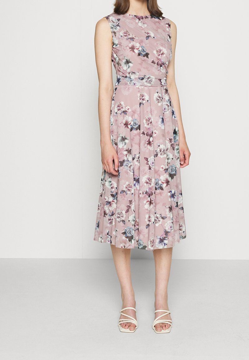 Femme vêtue d'une robe rose sans manches, longueur genou, à imprimé floral multicolore, et de sandales blanches à talons avec lanières, debout devant un fond uni.
