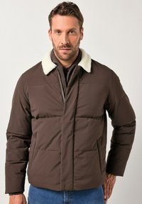 Man in een bruin pufferjack met witte sherpa kraag, gelaagd over een donkere zip-up sweater en blauwe jeans, staand tegen een effen achtergrond.