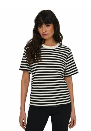 Donna con lunghi capelli scuri che indossa una maglietta a righe orizzontali bianco e nere a maniche corte e pantaloni neri, in piedi su sfondo bianco.