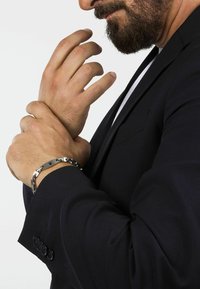 Bracciale in acciaio inossidabile con anelli intrecciati e finitura lucida, indossato su una mano con un giacca scura.