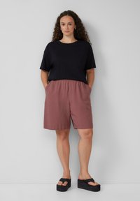 Schwarzes Baumwoll-T-Shirt in lockerer Passform, kombiniert mit burgunderroten Shorts mit elastischem Bund und seitlichen Taschen. Schwarze Plateausandalen.