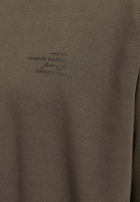 Ruda medvilnės marškiniai su švelnia tekstūra ir subtiliu žaliu logotipo spaudiniu, kuriame užrašyta "Premium Garment Supply" ir "Designed in Denmark."