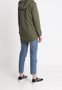 Olivgrön, långärmad parka med huva, med en rak passform, kombinerat med blå jeans och svarta sneakers med vita detaljer.