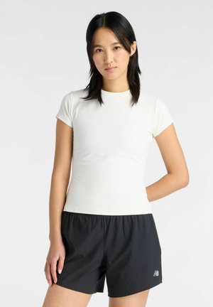 Nuori nainen, jolla on suorat mustat hiukset, pukeutuneena istuvaan valkoiseen lyhythihaiseen toppiin ja väljiin mustiin urheilushortseihin, seisoo yksinkertaista taustaa vasten.