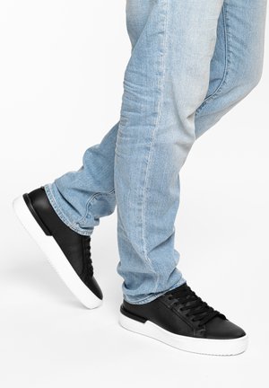 Beine in hellblauen Jeans und schwarzen Ledersneakern mit weißen Sohlen, ein Fuß leicht angehoben und hinter dem anderen überkreuzt.