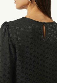 Schwarzes Oberteil mit strukturiertem Muster, Rundhalsausschnitt und Knopfverschluss im Rücken. Puffärmel verleihen dem Design Volumen.