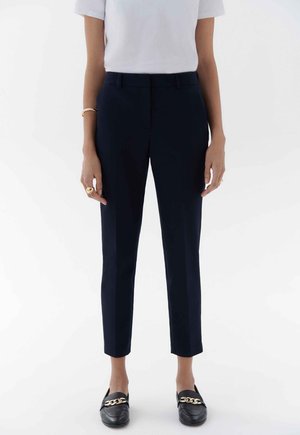 Pantalons ajustés bleu marine, avec une coupe slim, présentant un devant plat, des poches latérales invisibles et une texture lisse. Associés à des mocassins noirs.