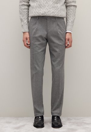 Uomo che indossa pantaloni grigi su misura, mocassini neri e un maglione a trecce di colore grigio chiaro, in piedi contro un muro beige.