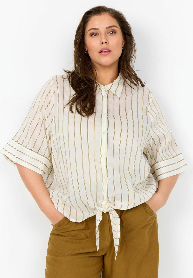Femme aux longs cheveux bruns portant une chemise blanche à manches courtes avec de fines rayures marron nouée à la taille et un pantalon marron, les mains dans les poches.
