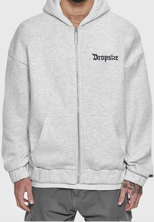 Homme portant un sweat à capuche gris clair zippé avec « Dropsize » en police gothique noire sur la poitrine gauche, mains tatouées et tatouage au cou « CHILD OF GOD ».