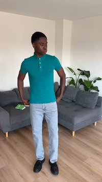 Νέος άνδρας με πετρόλ πόλο και ανοιχτόχρωμα τζιν στέκεται στο σαλόνι με γκρι καναπέ, πράσινο φυτό και ξύλινο δάπεδο.