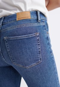 En smal passform blå denimjeans med en brun läder Superdry-logotyp på midjeresåren och en bakficka. Mjuk textur, vanlig tvätt.