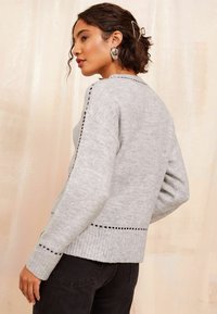 Grå strikket sweater med ribbet kant og prikkedetaljer langs ærmerne, iført med sorte bukser, mod en neutral baggrund.