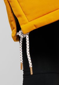 Tissu jaune à surface texturée, avec un cordon blanc orné d'accents marron à l'ourlet inférieur. Fond noir.