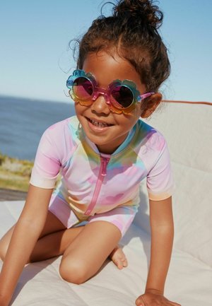 Junges Kind trägt bunte, blumenförmige Sonnenbrille und einen pastellfarbenen Tie-Dye-Reißverschluss-Einteil-Badeanzug, lächelt, während es draußen in der Nähe von Wasser sitzt.