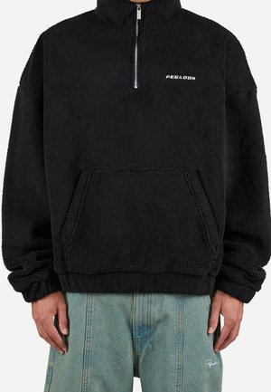 Pull en polaire noir avec fermeture éclair frontale et poche kangourou, porté avec un jean bleu clair, affichant un petit logo blanc "PEGADOR" sur la poitrine.