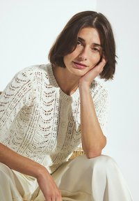 Una donna con capelli corti castani indossa un top a maglia bianco con motivo e pantaloni chiari, poggiando la testa sulla mano, seduta su una sedia di vimini.