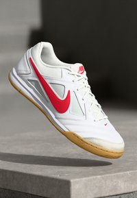 Nike sportiniai bateliai su balta sintetine viršutine dalimi, raudonu akcentu apnašu, šviesiai pilkomis tinklinėmis dalimis ir guminiu padu.