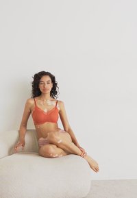Terrakottalace bralette og lyse rosa boyshorts båret av en sittende modell på en rund, teksturert beige sofa mot en hvit vegg.