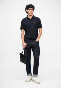 Námornícky polo tričko s bielymi akcentmi na golieri, spárované s tmavými džínsami. Model drží čiernu tašku na rameno a má na sebe biele tenisky.