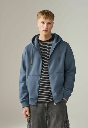 Noor mees lühikeste blondide juustega, kes kannab sinist lukuga kapuutsiga pusa musta-valgete triipudega t-särgi ja tumedate cargo-pükstega, seisab ühevärvilise tausta ees.