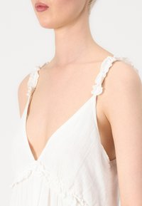 Hvit kjole med dyp V-hals, volanger på stroppene og strukturert stoff. Designet har flytende materiale og en elegant silhuett.