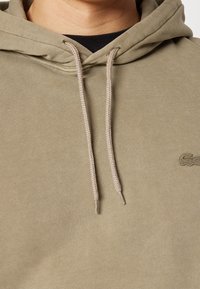Hoodie verde oliva chiaro con tessuto testurizzato, stringhe in un beige più chiaro e un piccolo logo sul lato sinistro anteriore.