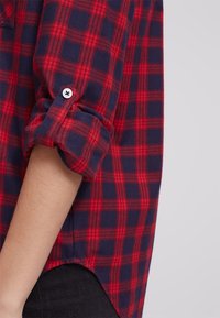 Geruite shirt in rood en marineblauw, gemaakt van zachte stof. Kenmerkt zich door opgerolde mouwen en een gebogen zoom. Zichtbaar knoopdetail op de mouw.
