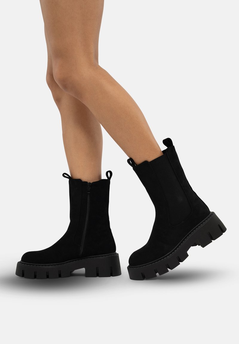 Black suede ankle boots with a chunky rubber sole, featuring a side zipper and pull tabs. Smooth texture and minimalistic design.