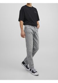 Hombre vistiendo una camiseta negra de gran tamaño, jeans gris claro y zapatillas altas negras caminando contra un fondo liso gris claro.