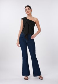 Μαύρο τοπ με έναν ώμο και δέσιμο κόμβου, συνδυασμένο με σκουρό μπλε καμπάνα τζιν και μαύρα σανδάλια με λουράκια. Λείο ύφασμα και εφαρμοστή σιλουέτα.