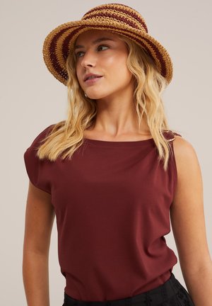 Plavokosa žena koja nosi prugasti slameni šešir i bordo top bez rukava, gleda blago u stranu na jednobojnoj pozadini.