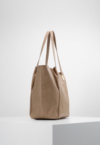 Bolso tote de cuero beige con un diseño flexible, base plana y dos asas largas. Textura suave con un pequeño acento metálico.