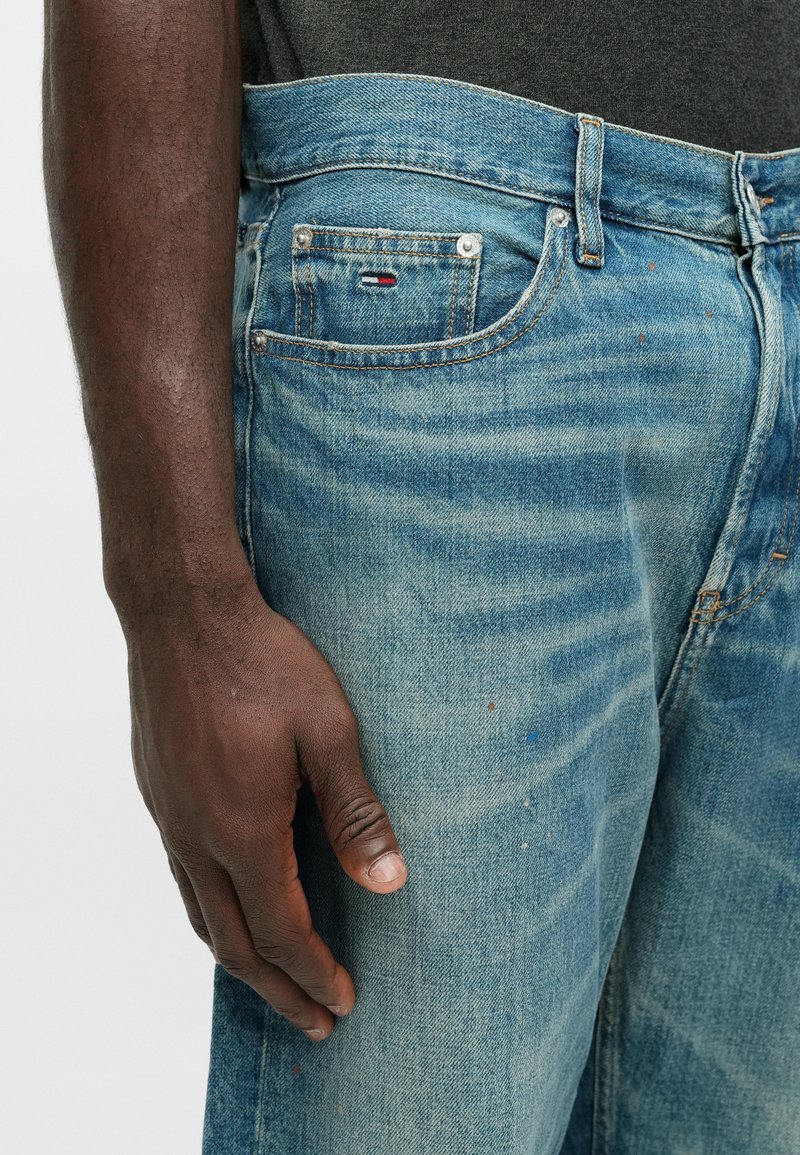 Light blue denim jeans with a faded design, a small front pocket, and contrasting stitching. Visible wear and small colorful specks.