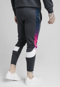 Dunkelgraue, verjüngte Jogginghosen mit einem Verlaufsseitenstreifen in Blau, Pink und weißen Akzenten, mit gerippten Bündchen und Taschen. Kombiniert mit weißen Sneakers.