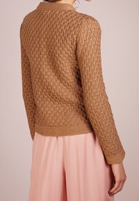 Maglione lavorato a maglia in un caldo colore marrone con un motivo a diamante testurizzato, scollo rotondo e polsini e bordo a costine. Indossato sopra un indumento rosa chiaro.