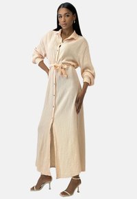 Lang knap-op kjole i ferskenfarve med tekstureret stof og bindebælte i taljen. Har opsmøgede ærmer og en frontslids.