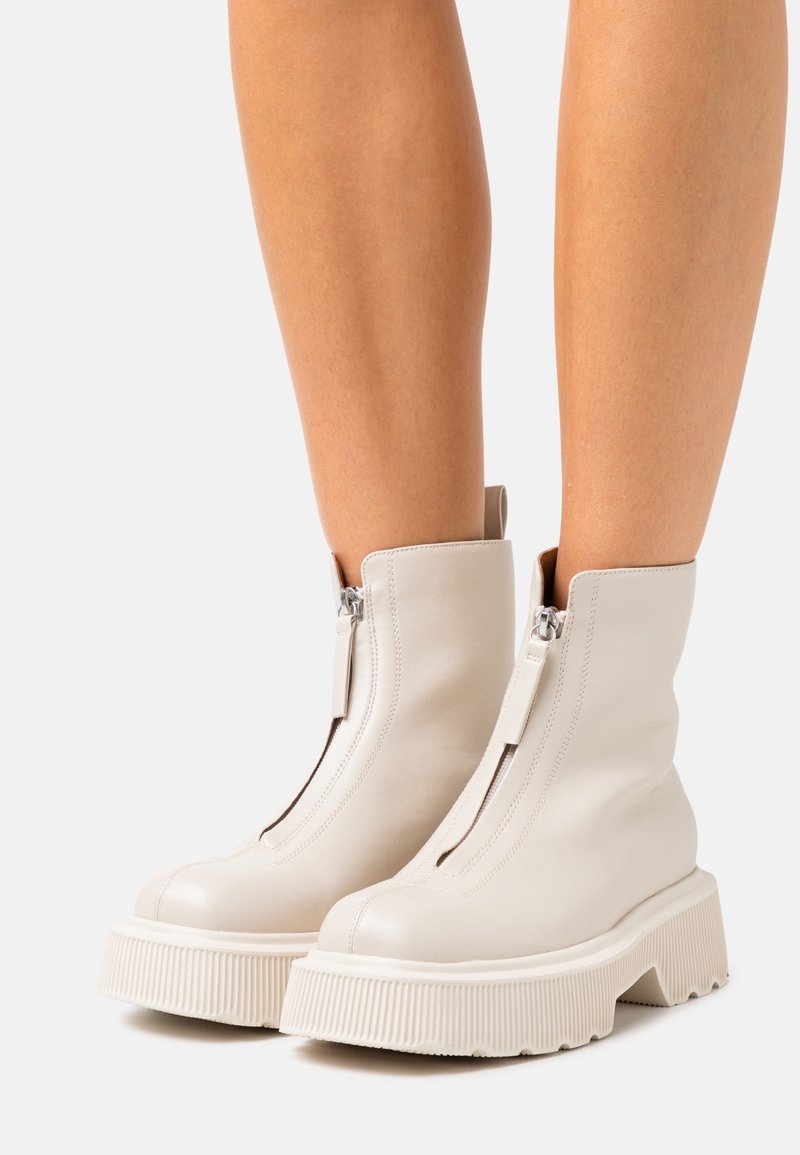 Botas de tobillo de cuero blanco con suela de goma gruesa, que cuentan con una cremallera frontal y detalles acanalados en la suela. Textura suave y diseño sencillo.