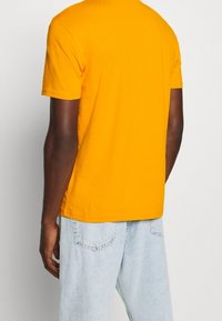 Žluté tričko s krátkým rukávem z bavlny, s kulatým výstřihem, uvolněného střihu a jednoduchým designem na zadní straně. V kombinaci se světle modrými džíny.