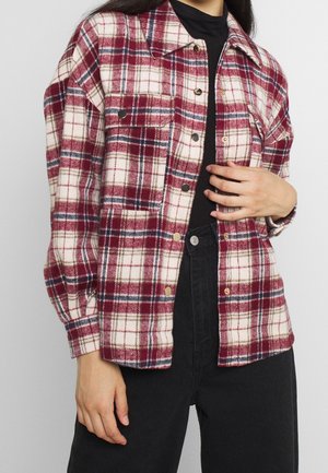 Personne portant une chemise à carreaux rouge, blanche et bleu marine boutonnée par-dessus un haut noir et un pantalon noir, la main reposant sur le ventre.